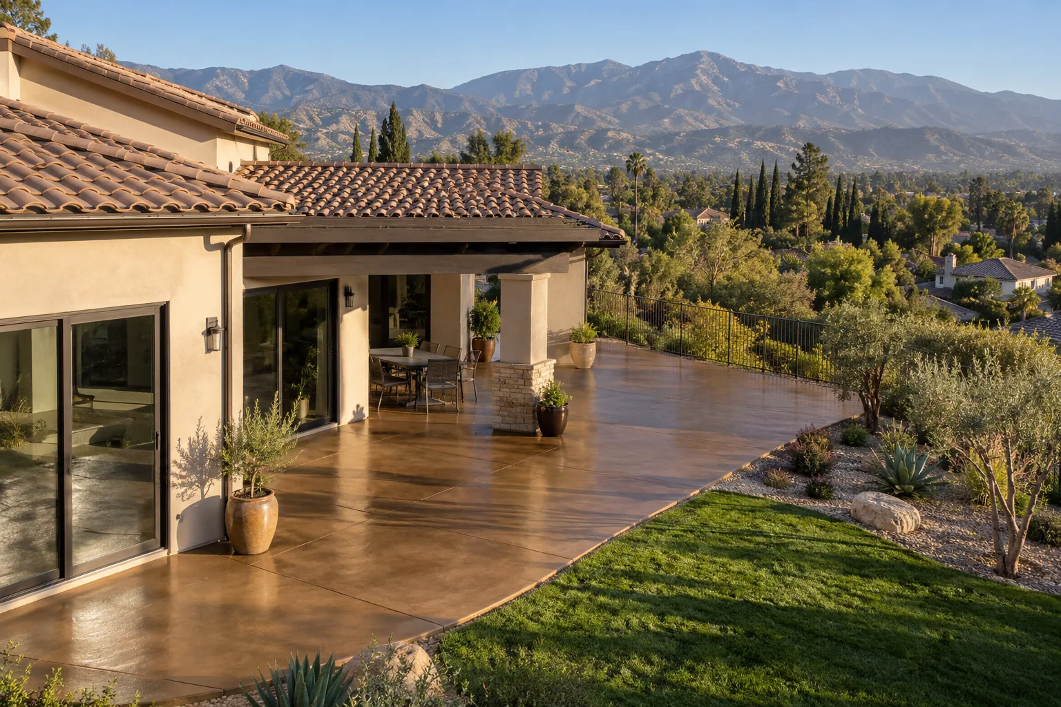Back patio in warm two-tone earth-tone stained and sealed concrete with potted plants, a lawn and mountains beyond