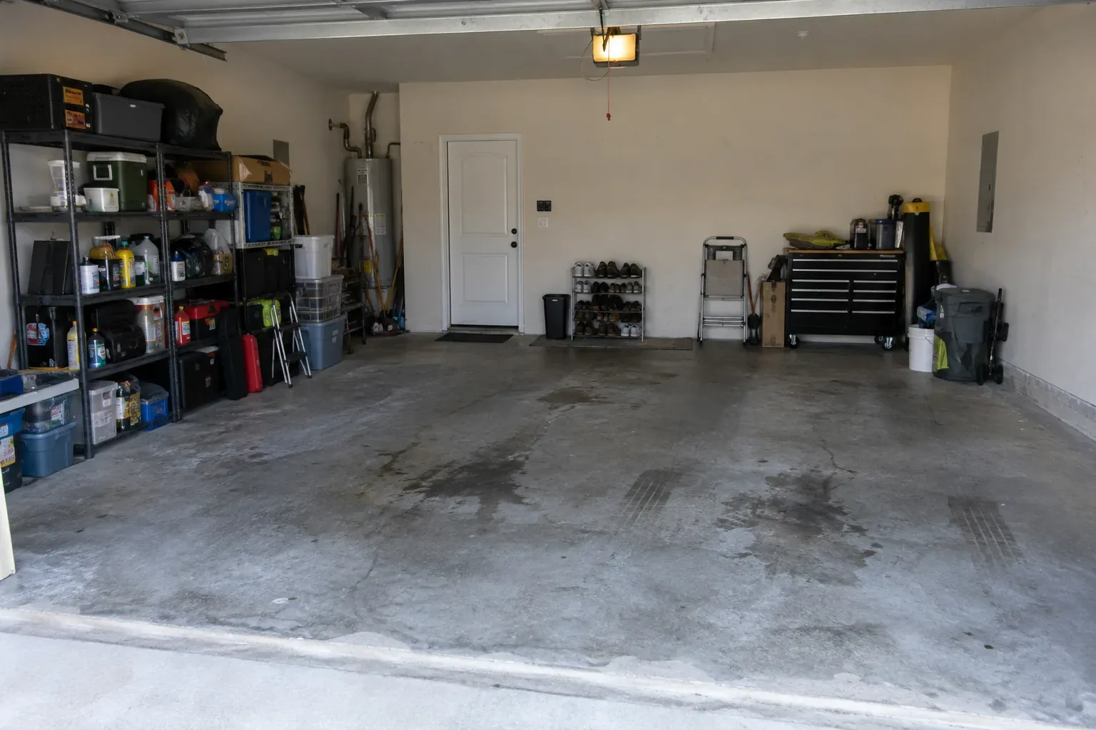 Riverside garage before epoxy: a bare two-car slab with oil stains and worn patches, metal shelving with storage bins on the left and a tool chest at the back